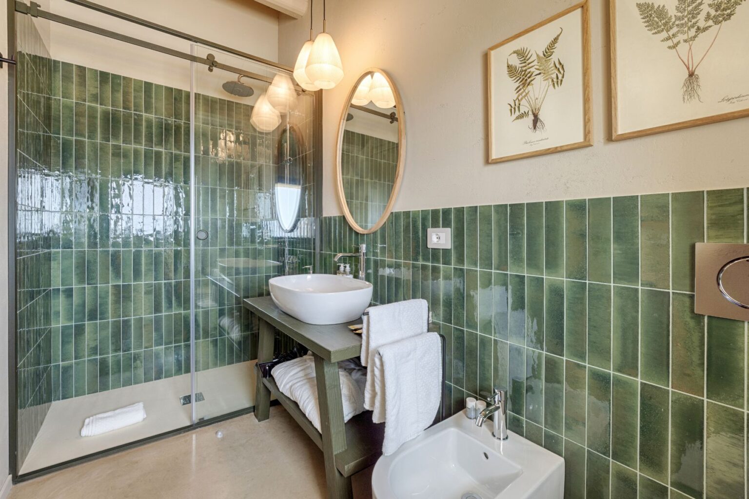 Modern bathroom featuring a glass shower, green tiled walls, a round mirror, and a stylish sink on a wooden vanity. Soft towels are neatly arranged, complemented by minimalist decor and natural lighting. | Only on SopranoVillas