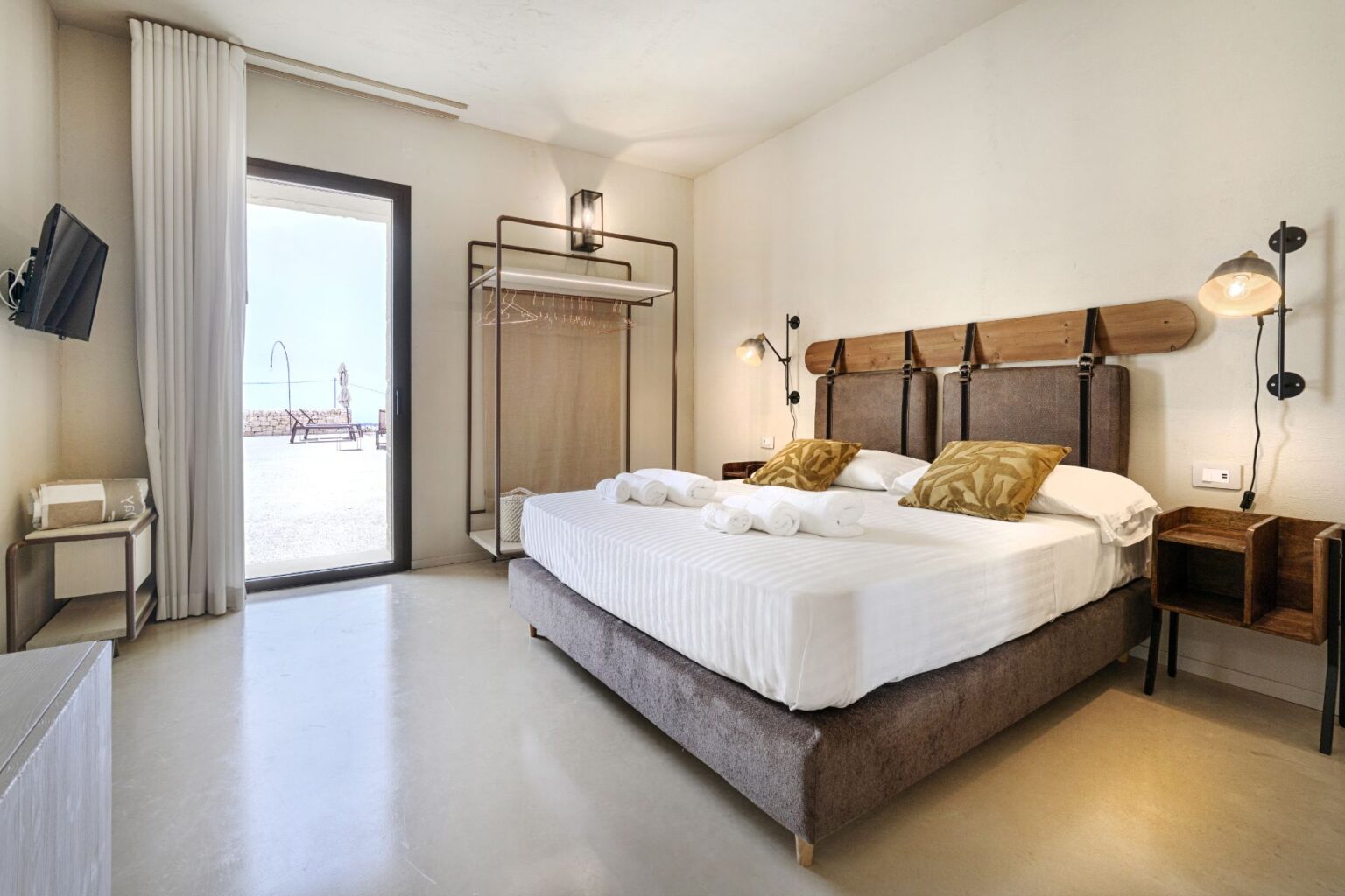 Modern hotel bedroom with a double bed, decorative pillows, and neatly arranged towels. Features a minimalist design with a mounted TV, stylish lamps, and an open closet. Large glass door leads to a sunny outdoor area with lounge chairs. Ideal for a relaxing getaway. | Only on SopranoVillas