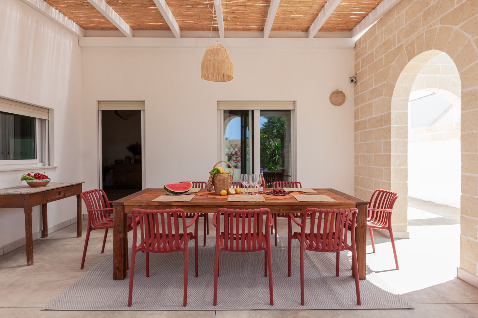 Outdoor dining area featuring a wooden table surrounded by red chairs, adorned with fresh fruits and a woven basket, set against a backdrop of white walls and natural stone accents. | Only on SopranoVillas