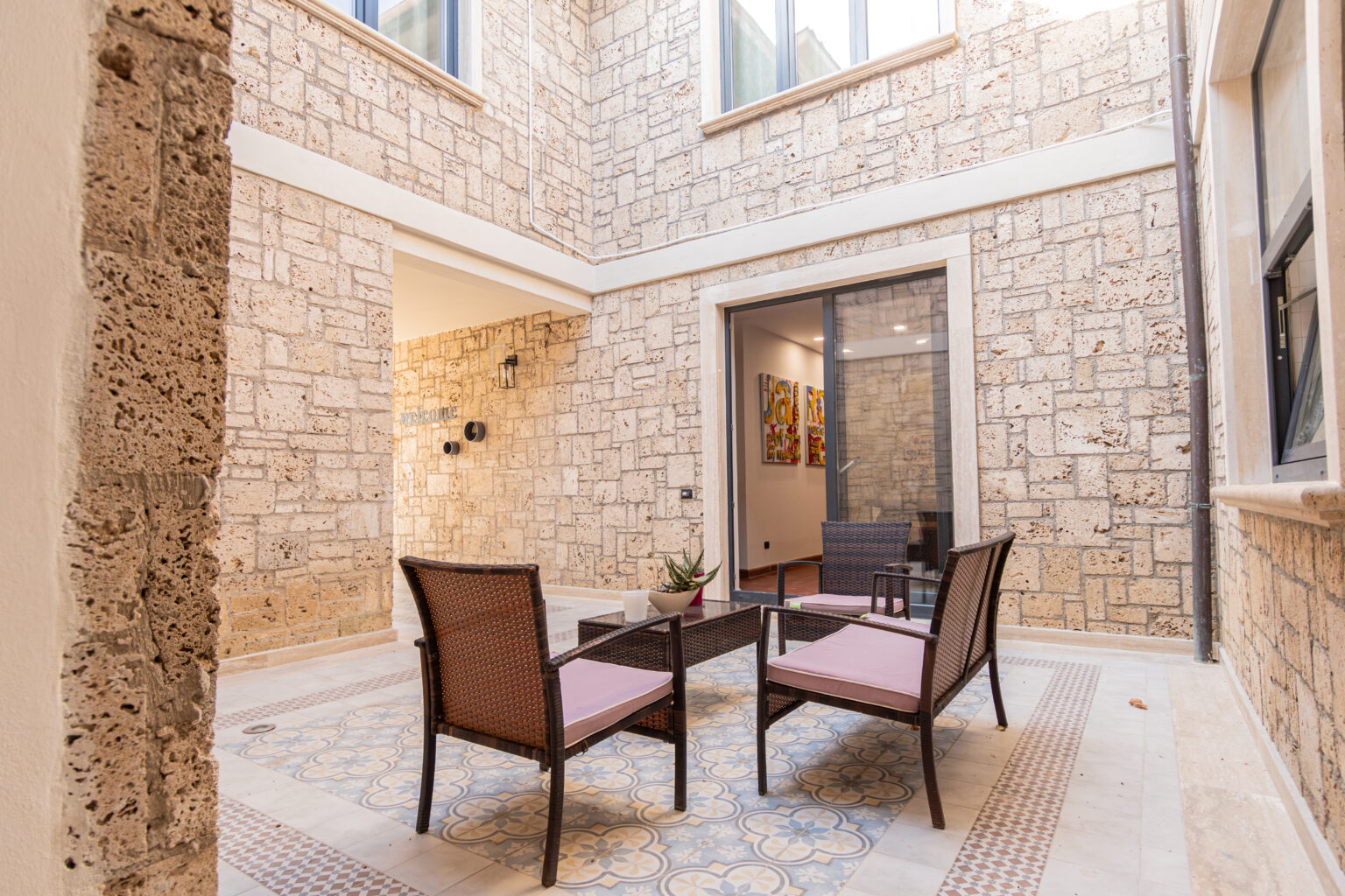 Courtyard with modern seating featuring two wicker chairs and a coffee table, surrounded by textured stone walls and decorative tiled flooring, offering a serene outdoor space. | Only on SopranoVillas