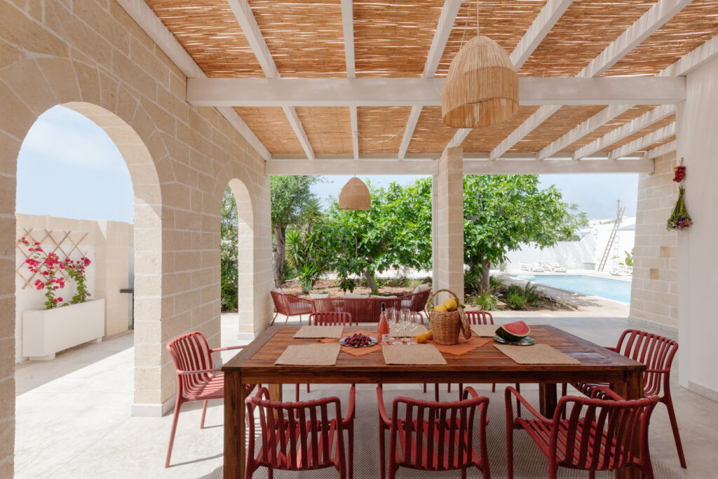 Outdoor dining area featuring a wooden table surrounded by red chairs, adorned with fresh fruits and glasses, under a straw-covered roof. Lush greenery and a swimming pool are visible in the background, creating a serene and inviting atmosphere. | Only on SopranoVillas