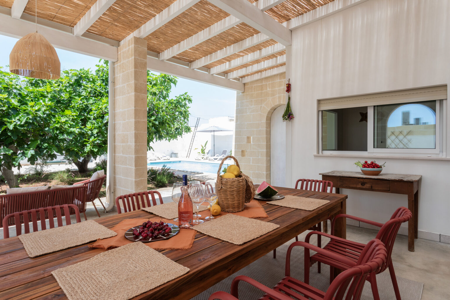 Outdoor dining area featuring a wooden table set with placemats, a basket of fruits, and a bottle of rosé wine. The space is shaded by a straw roof and surrounded by lush greenery, with a glimpse of a swimming pool in the background. | Only on SopranoVillas