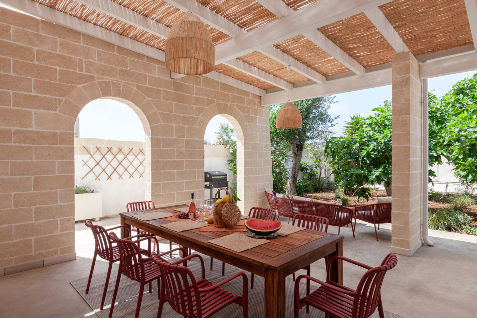 Outdoor dining area featuring a wooden table with red chairs, surrounded by natural stone walls and arches. The space is adorned with hanging light fixtures and includes a barbecue grill, lush greenery, and decorative fruit arrangements, creating a cozy and inviting atmosphere for gatherings. | Only on SopranoVillas