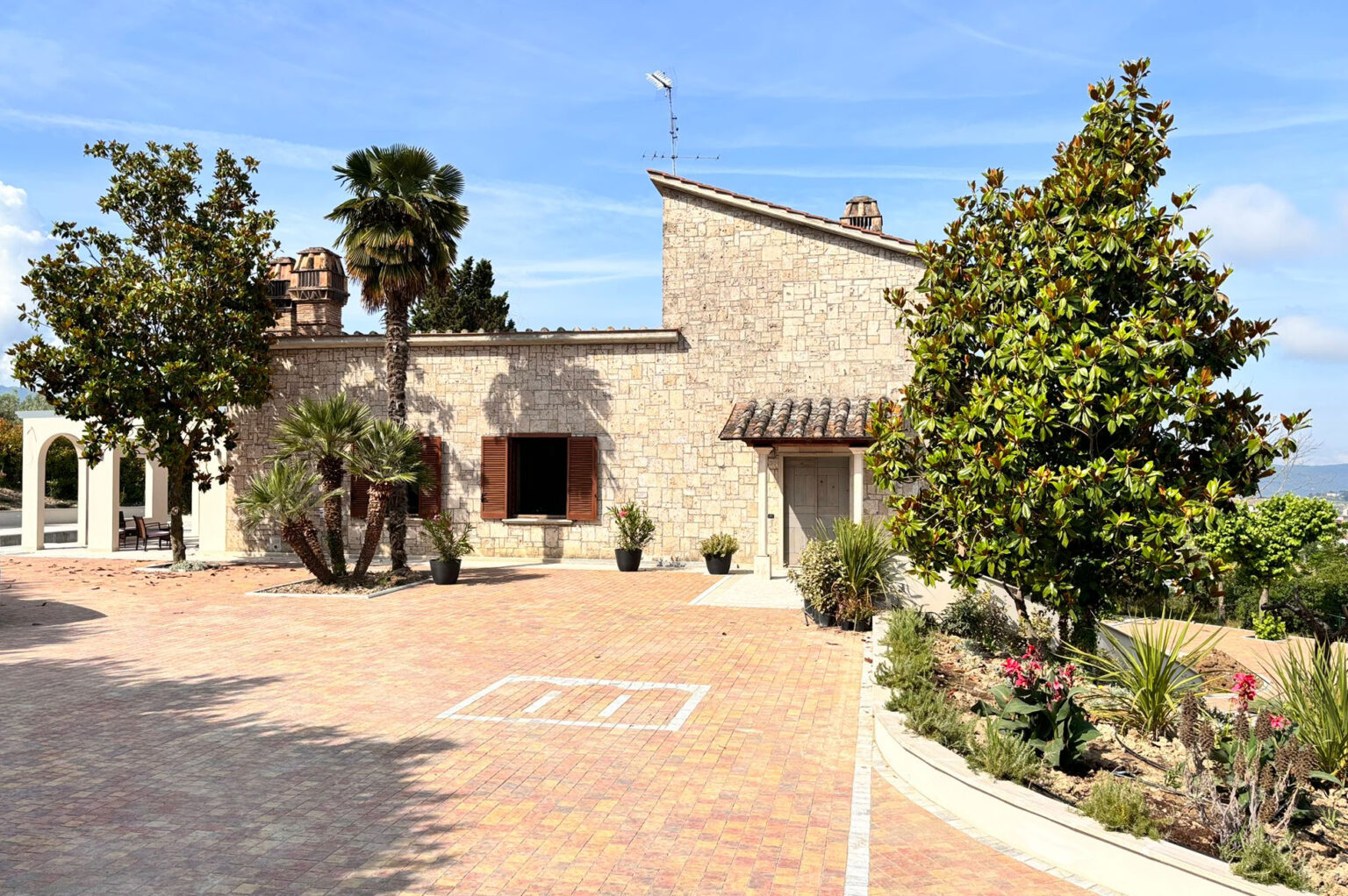 Charming stone house surrounded by lush greenery and palm trees, featuring a paved driveway and decorative landscaping, under a clear blue sky. | Only on SopranoVillas