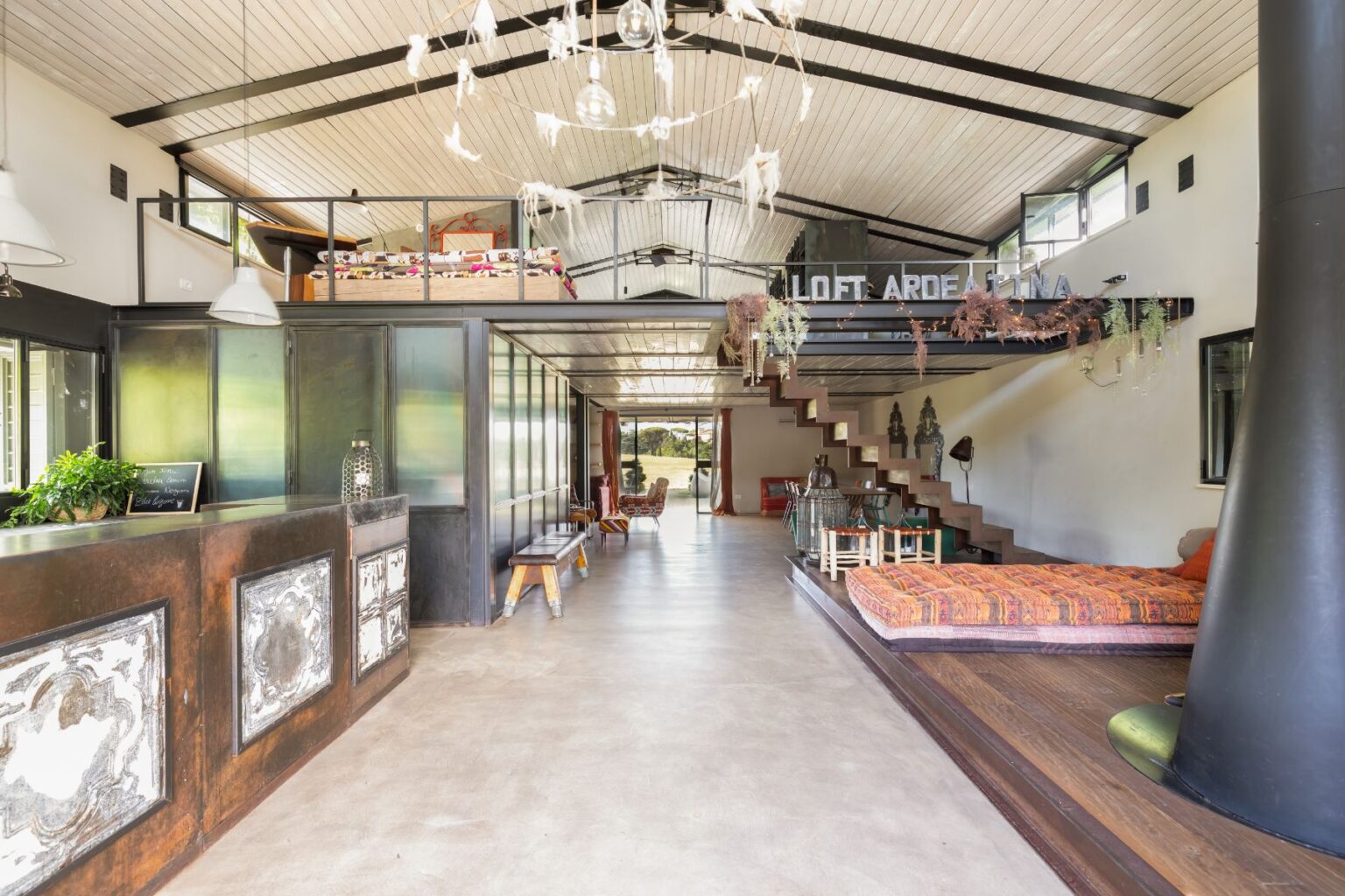 Modern loft interior featuring a spacious open layout with high ceilings, a stylish staircase, and eclectic furnishings. The design includes a reception area with a decorative counter, vibrant seating, and large windows that offer natural light and views of the outdoors. Ideal for showcasing contemporary living spaces. | Only on SopranoVillas