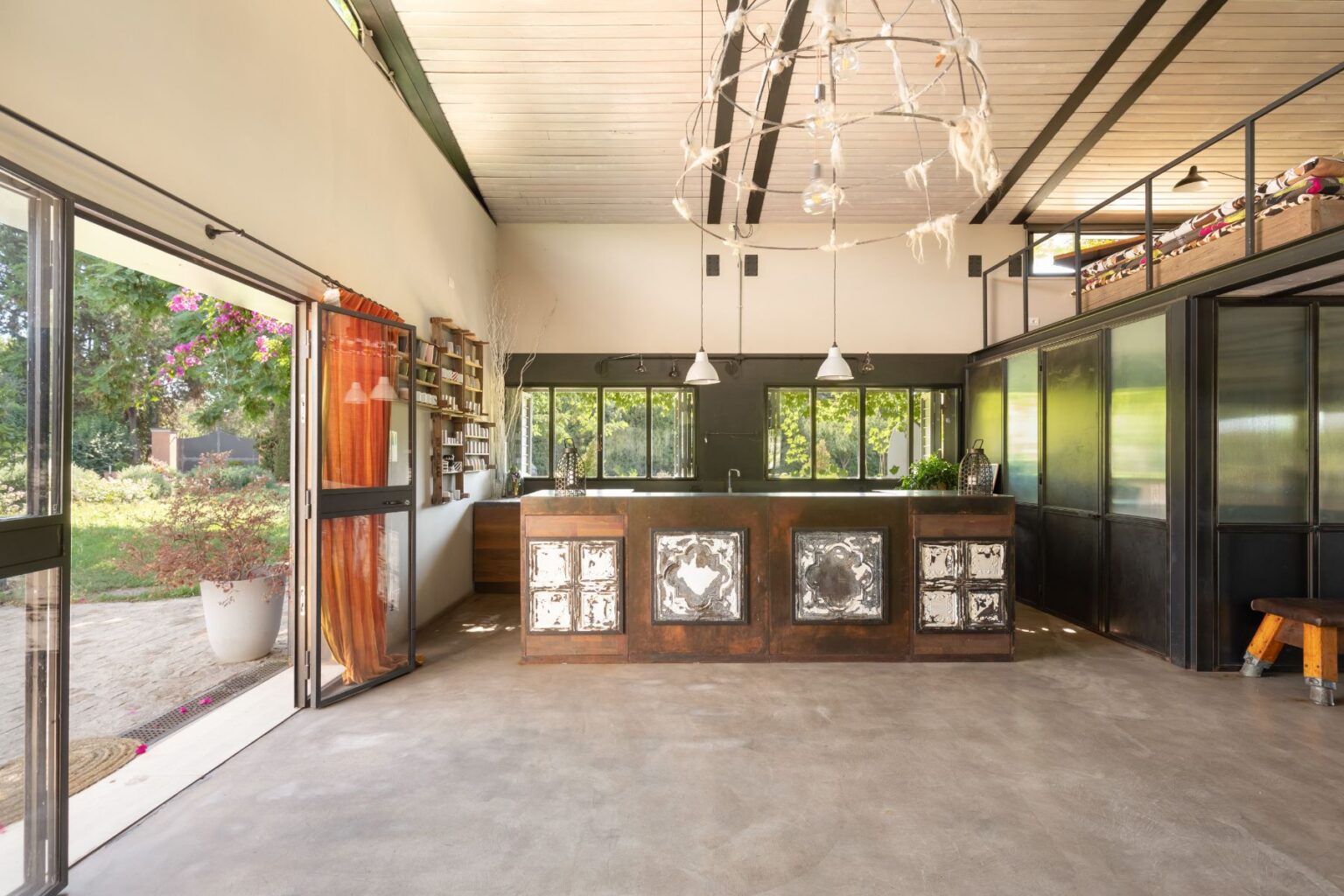 Modern kitchen interior featuring a spacious layout with a rustic metal island, large windows, and natural light. The design includes wooden accents, a unique chandelier, and a cozy atmosphere, ideal for contemporary living. Bright greenery is visible outside through open doors. | Only on SopranoVillas