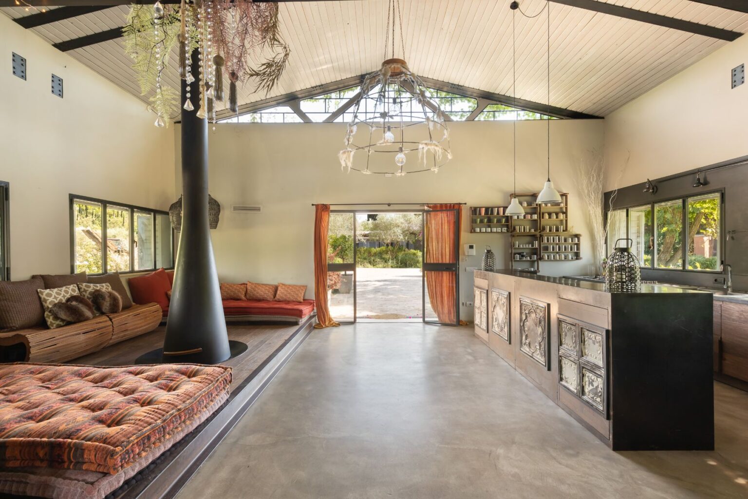 Modern open-concept living space featuring a cozy seating area with colorful cushions, a sleek kitchen with decorative elements, and large windows that provide natural light and a view of the garden outside. The design includes a unique black chimney and an artistic chandelier, creating a warm and inviting atmosphere. | Only on SopranoVillas