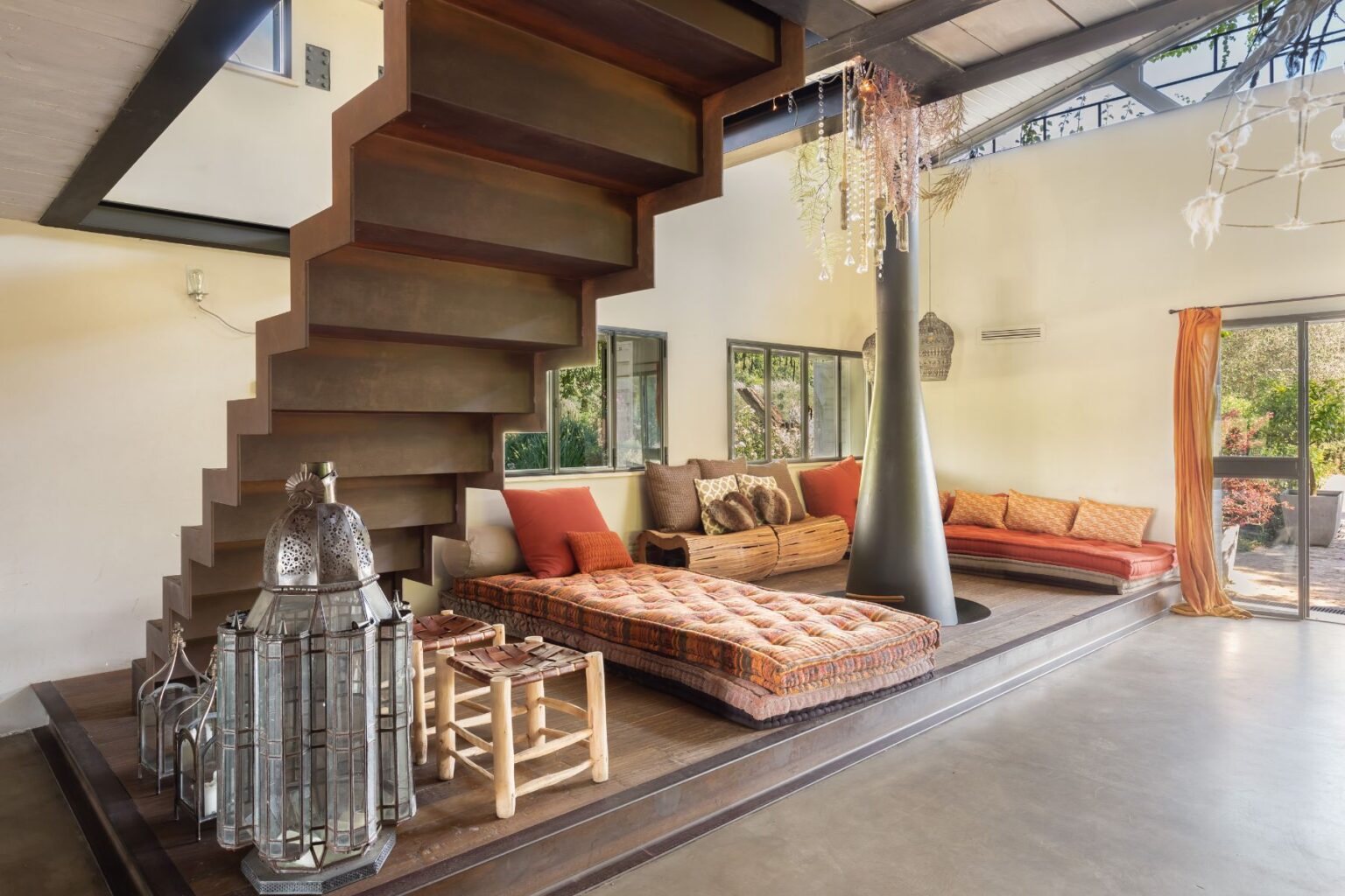 Modern living room featuring a stylish staircase, cozy seating areas with cushions, and decorative lanterns. Natural light floods the space through large windows, highlighting the warm earthy tones and unique design elements. | Only on SopranoVillas