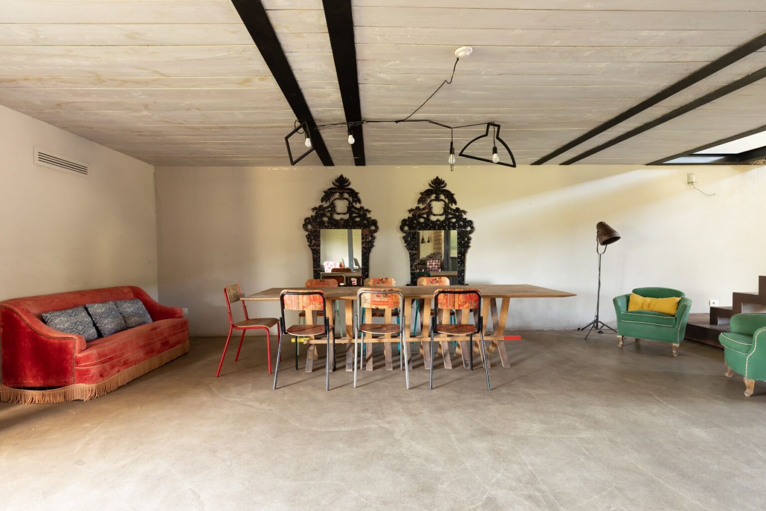 Modern dining room featuring a wooden table surrounded by colorful chairs, a plush red sofa, and two ornate mirrors on the wall. The space includes stylish lighting and a green accent chair, showcasing a blend of contemporary and vintage decor. | Only on SopranoVillas