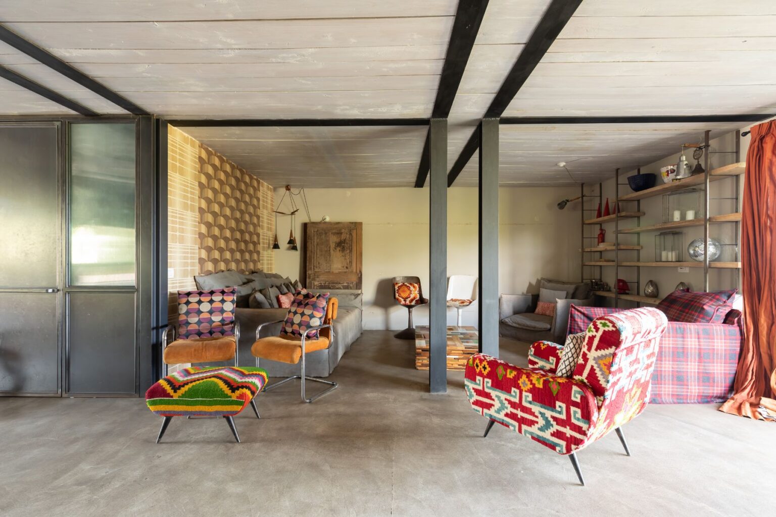 Stylish modern living room featuring colorful patterned chairs, a cozy sofa, and a mix of textures. The space showcases a contemporary design with industrial elements, decorative wall patterns, and a warm color palette, creating an inviting atmosphere for relaxation and socializing. | Only on SopranoVillas