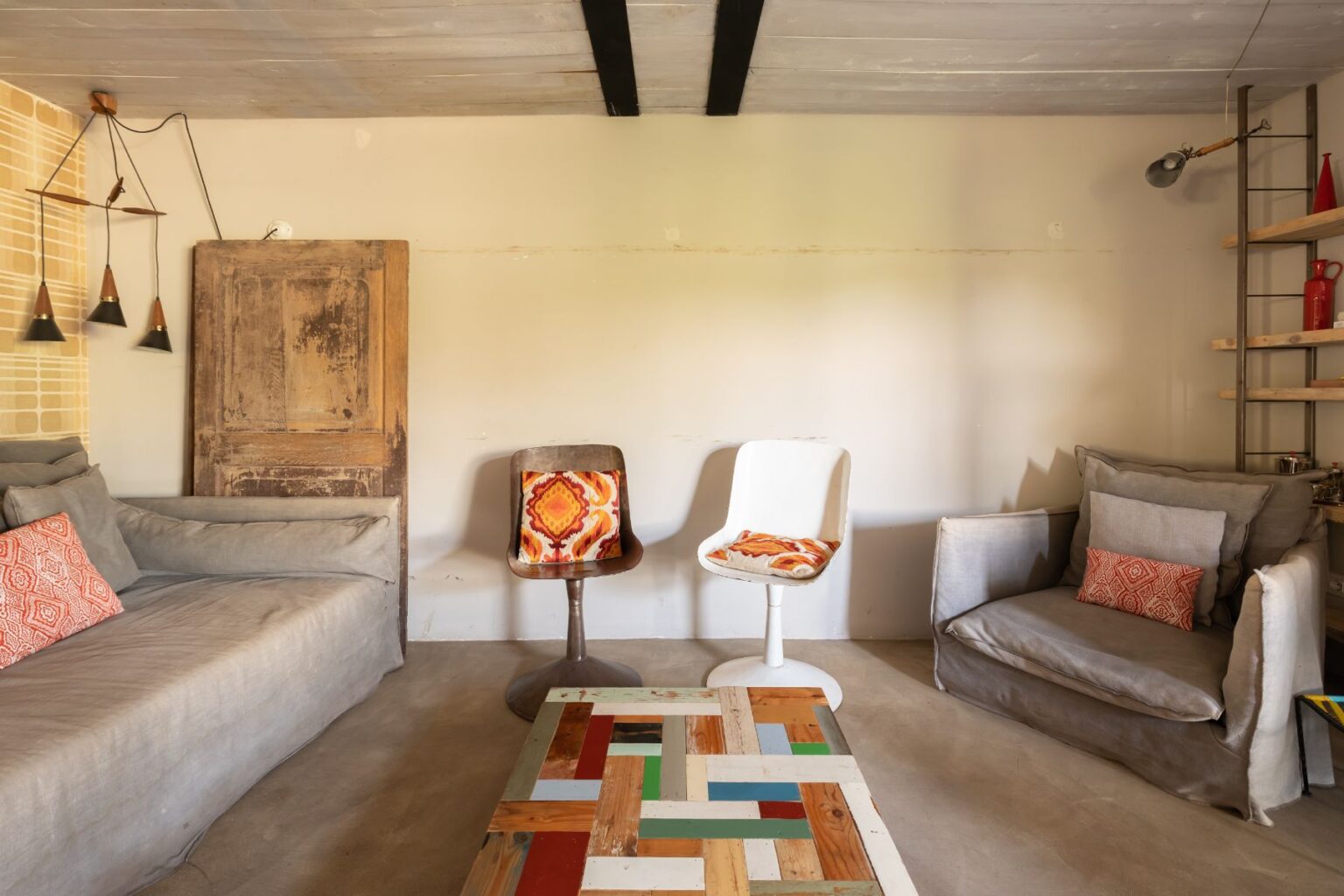 Cozy living room featuring a gray sofa with decorative pillows, two stylish chairs with patterned cushions, a colorful wooden coffee table, and a rustic wooden door. Ambient lighting is provided by modern pendant lights hanging from the ceiling, creating a warm and inviting atmosphere. | Only on SopranoVillas
