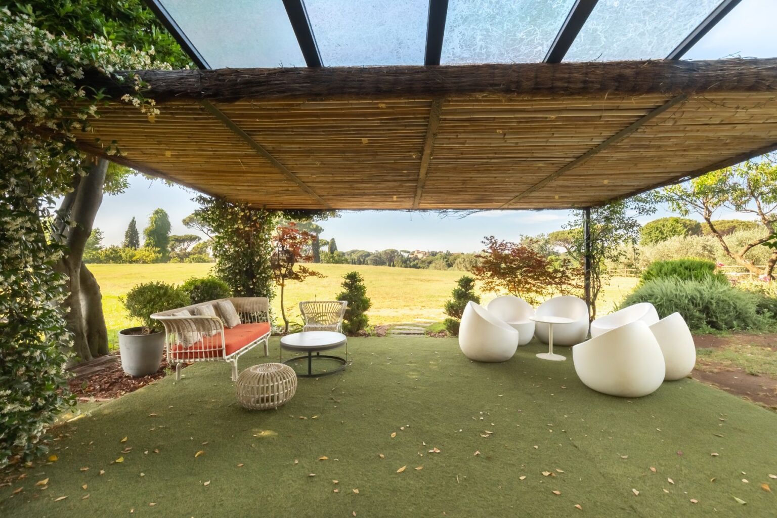 Modern outdoor lounge area with stylish white seating, a round coffee table, and a cozy sofa with orange cushions, surrounded by lush greenery and a scenic view of a field under a bamboo canopy. Ideal for relaxation and entertaining. | Only on SopranoVillas
