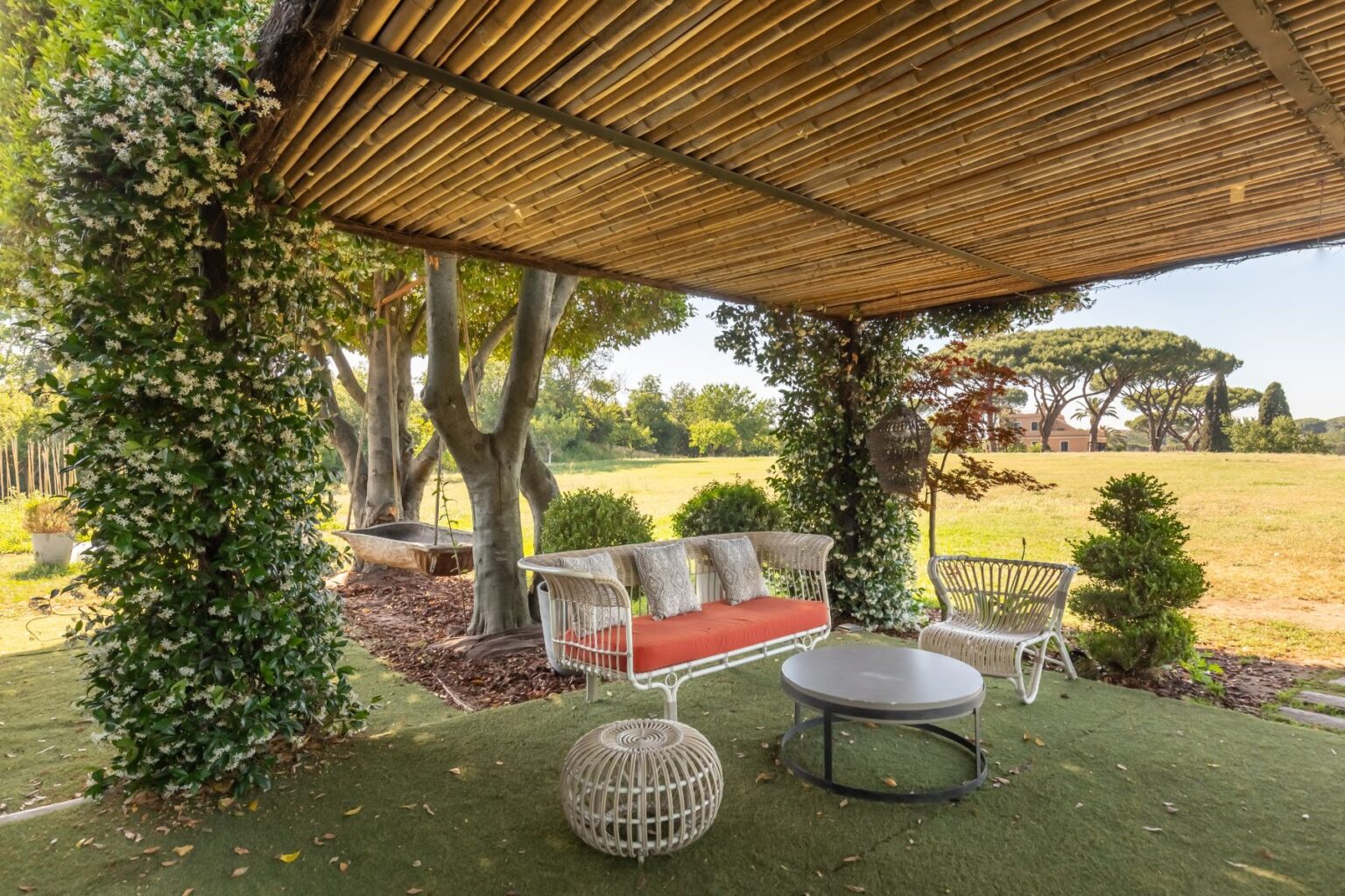 Cozy outdoor seating area under a bamboo roof, featuring a white wicker couch with orange cushions, a round coffee table, and lush greenery, set against a serene landscape with trees and open fields. Ideal for relaxation and outdoor gatherings. | Only on SopranoVillas