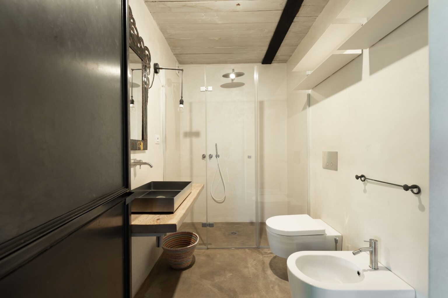 Modern bathroom featuring a glass shower, minimalist black sink, wooden countertop, and sleek white fixtures. The space is illuminated with stylish pendant lights and has a contemporary design with a concrete floor and decorative mirror. | Only on SopranoVillas