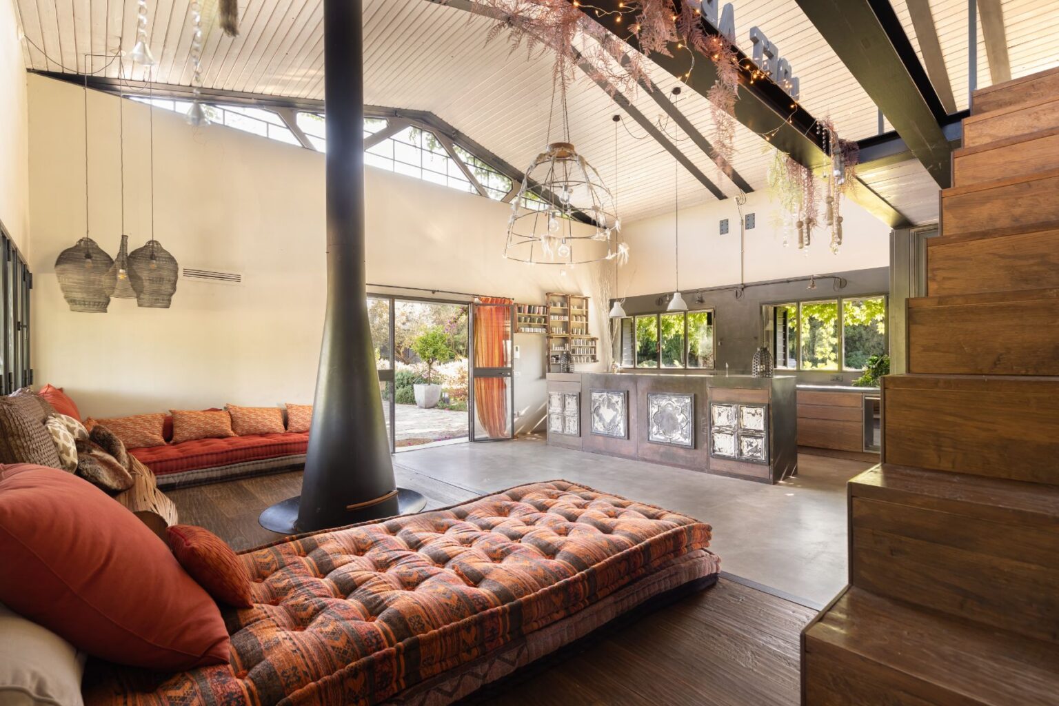 Modern open-concept living room featuring a cozy seating area with colorful cushions, a sleek black wood-burning stove, and a stylish kitchen with metallic accents. Large windows provide ample natural light and a view of the outdoor garden. Unique decorative elements include pendant lighting and rustic wooden stairs. | Only on SopranoVillas