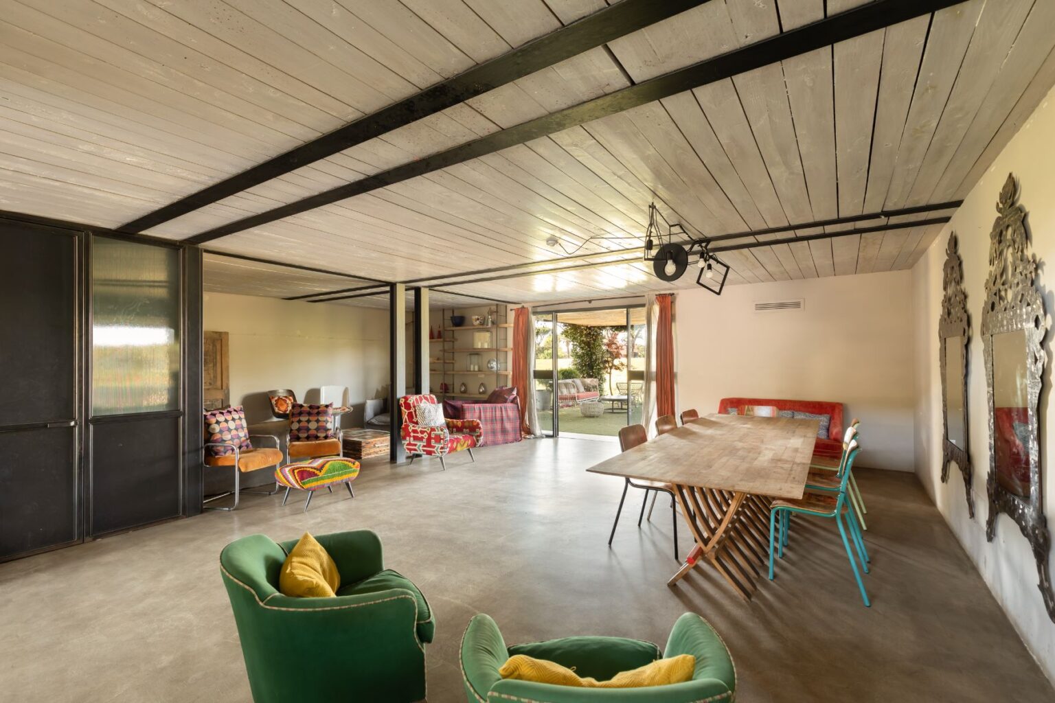Spacious modern living area featuring a large wooden dining table, colorful chairs, and vibrant decor. The room has large windows that open to a garden view, with a mix of contemporary and eclectic furniture styles, including green armchairs and patterned seating. Ideal for social gatherings and family meals. | Only on SopranoVillas