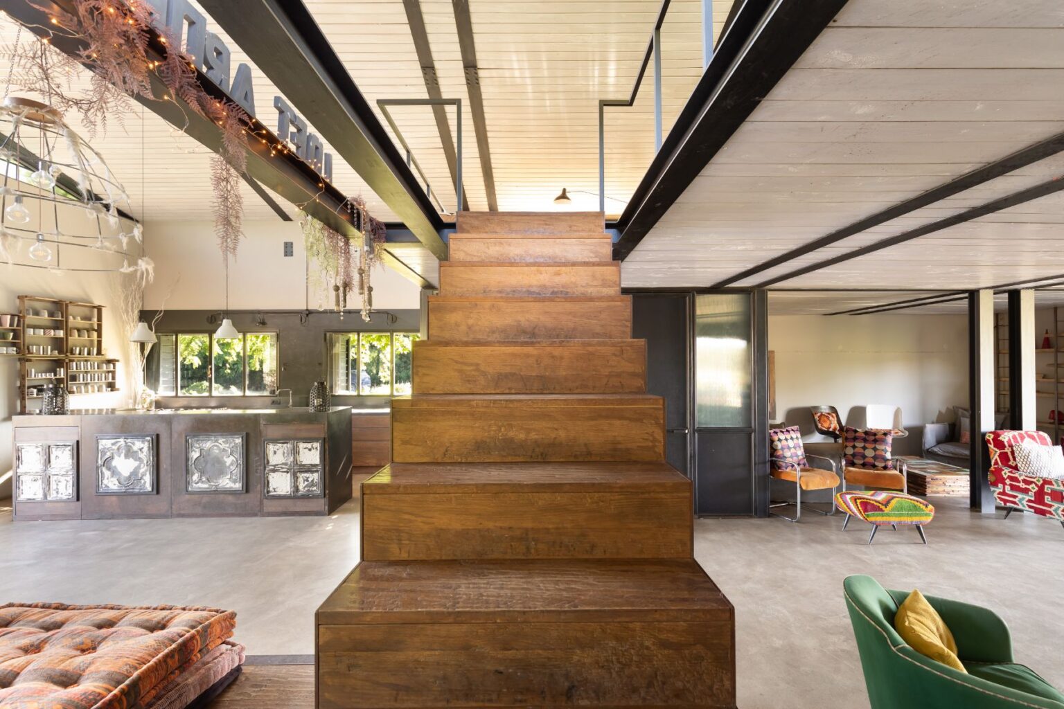 Modern interior design featuring a wooden staircase leading to an upper level, with a stylish living area and kitchen in the background. The space showcases contemporary furnishings, large windows for natural light, and decorative elements like plants and unique light fixtures. | Only on SopranoVillas