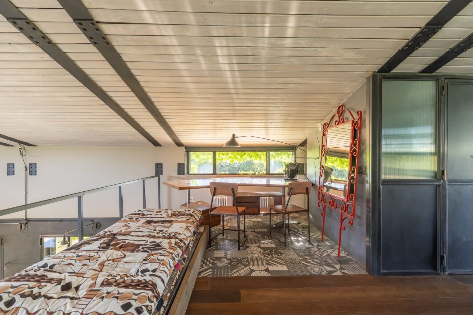 Cozy loft interior featuring a bed with a patterned quilt, a small dining area with wooden chairs, and large windows providing natural light. The space showcases modern design elements, including a decorative red mirror and patterned flooring, creating a stylish and inviting atmosphere. | Only on SopranoVillas