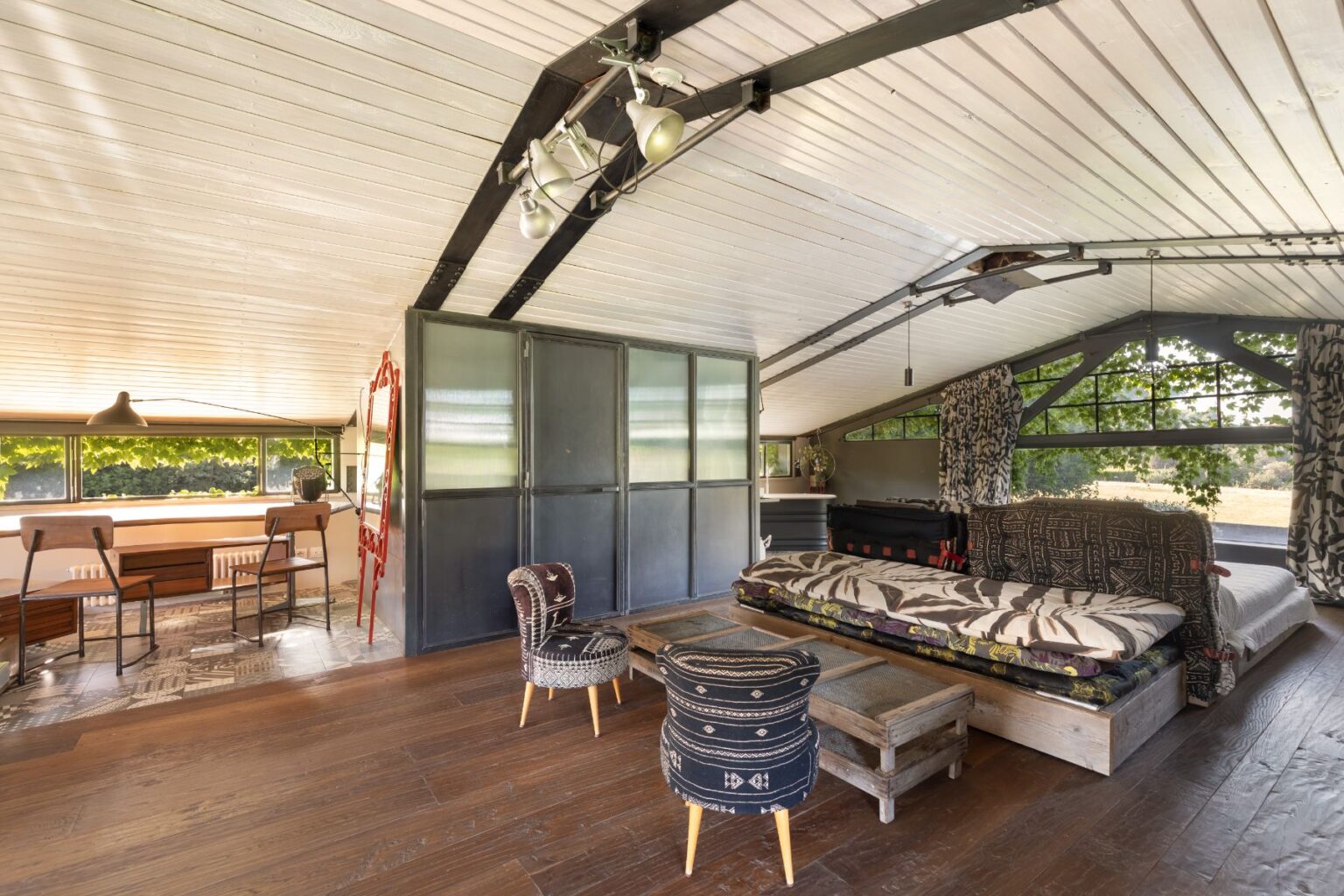Stylish loft interior featuring a wooden floor, large windows with natural light, a cozy bed adorned with patterned bedding, a rustic coffee table, and contemporary seating arrangements, ideal for modern living and relaxation. | Only on SopranoVillas