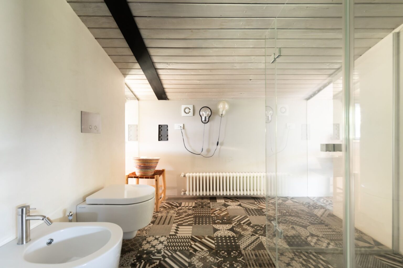 Modern bathroom interior featuring a minimalist design with a bidet and toilet, stylish patterned flooring, and unique light fixtures. The space is bright and airy, enhanced by glass shower walls and a wooden accent table. | Only on SopranoVillas