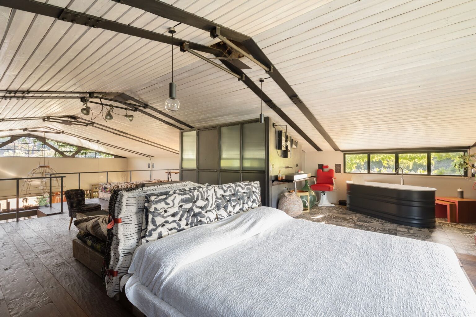 Spacious and modern loft bedroom with a white quilted bed, featuring a stylish headboard and wooden flooring. The room includes a cozy seating area, a sleek black bathtub, and large windows allowing natural light. Industrial-style beams and contemporary decor create a chic atmosphere. | Only on SopranoVillas