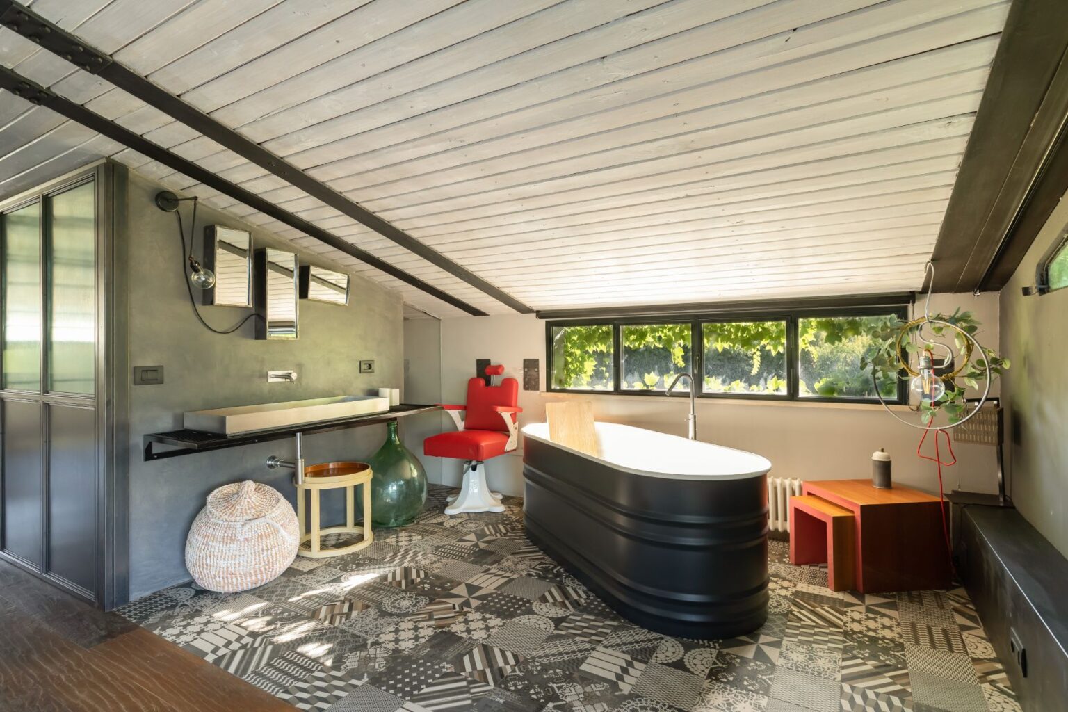 Modern interior design featuring a stylish bathroom with a freestanding black bathtub, a red accent chair, and a unique patterned floor. The space is illuminated by natural light through large windows, showcasing a minimalist aesthetic with a blend of textures and colors. | Only on SopranoVillas