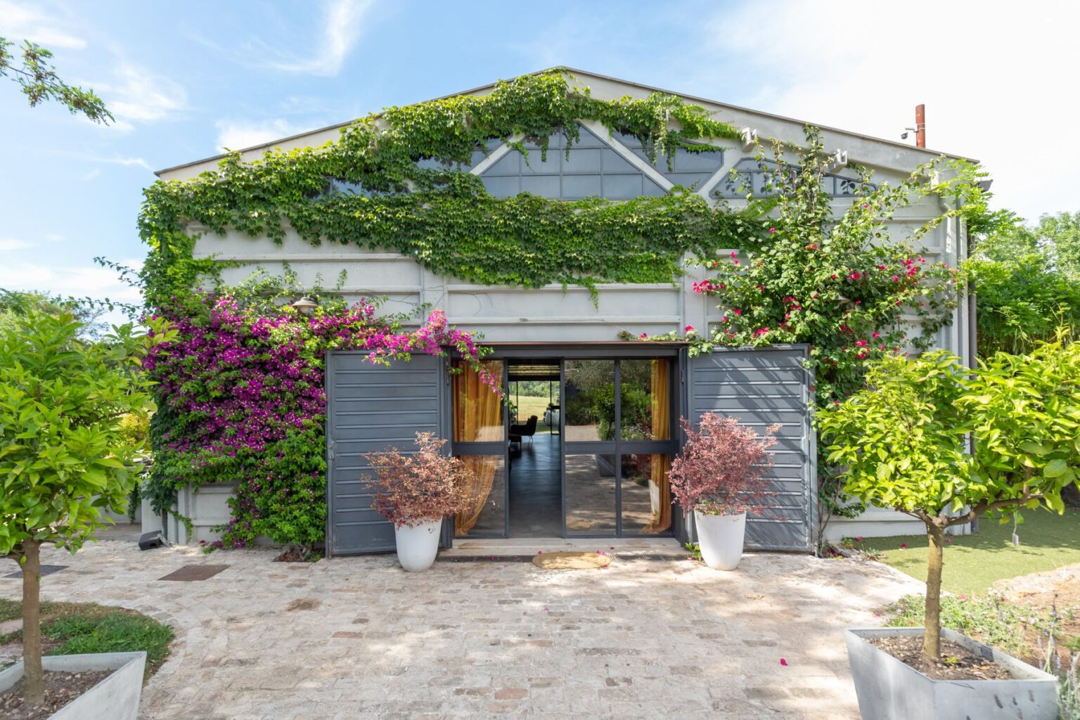 Modern architectural design featuring a spacious entrance adorned with vibrant greenery and colorful flowers. The building's exterior showcases large windows and double doors, inviting natural light into the interior. Well-maintained garden elements, including potted plants and a stone pathway, enhance the welcoming atmosphere of this stylish home. | Only on SopranoVillas