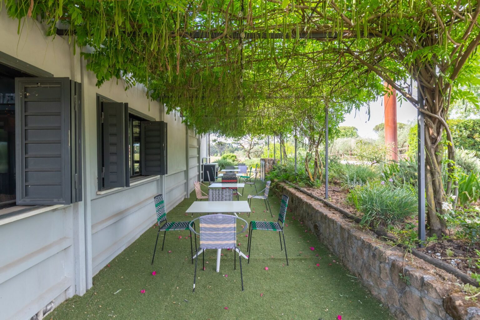 Outdoor dining area with tables and colorful chairs under a lush green canopy, featuring a modern building facade and landscaped garden. Ideal for enjoying meals in a serene setting. | Only on SopranoVillas