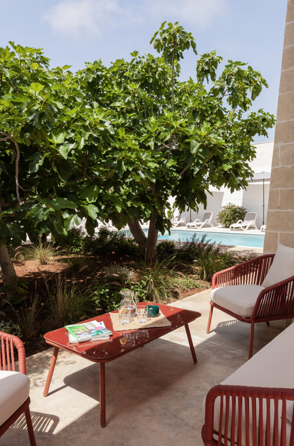 Cozy outdoor patio featuring a red table set with a pitcher and glasses, surrounded by lush green foliage and a view of a swimming pool in the background. Ideal for relaxation and leisure. | Only on SopranoVillas