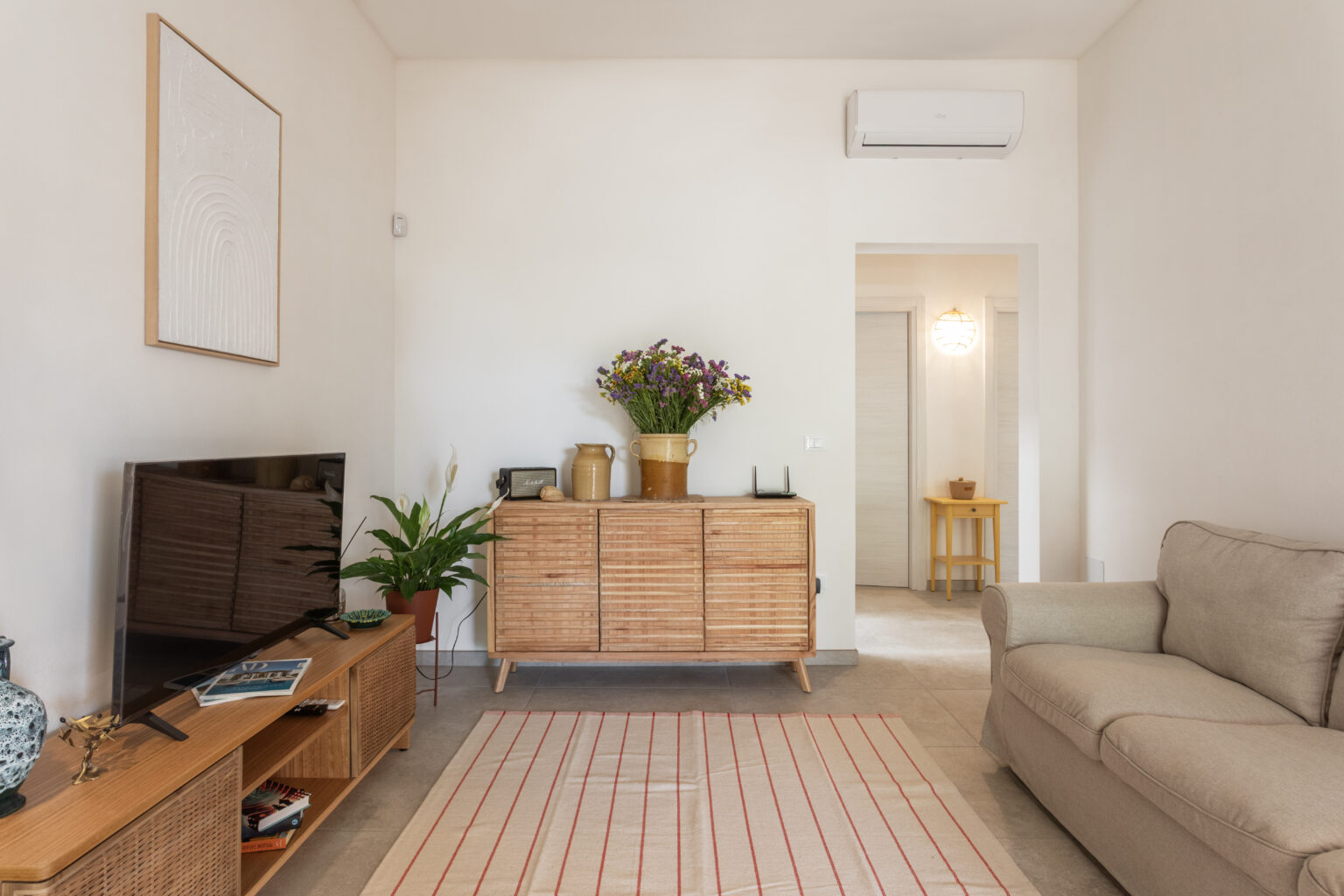 Bright and modern living room featuring a beige sofa, a stylish wooden sideboard adorned with a vase of flowers, and a flat-screen TV. A striped rug adds texture to the space, complemented by indoor plants and a glimpse of a cozy dining area in the background. | Only on SopranoVillas