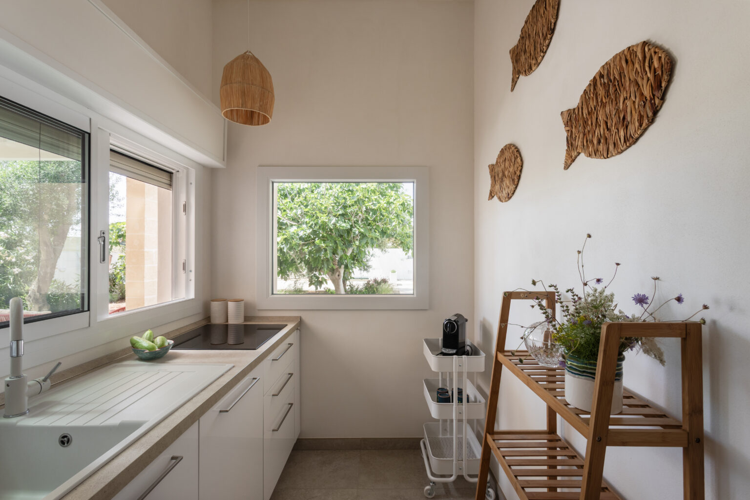Bright and modern kitchen featuring a sink, countertop, and stylish decor. A large window offers a view of greenery outside, while decorative fish wall art and a wooden shelf with plants add a touch of warmth and character. | Only on SopranoVillas