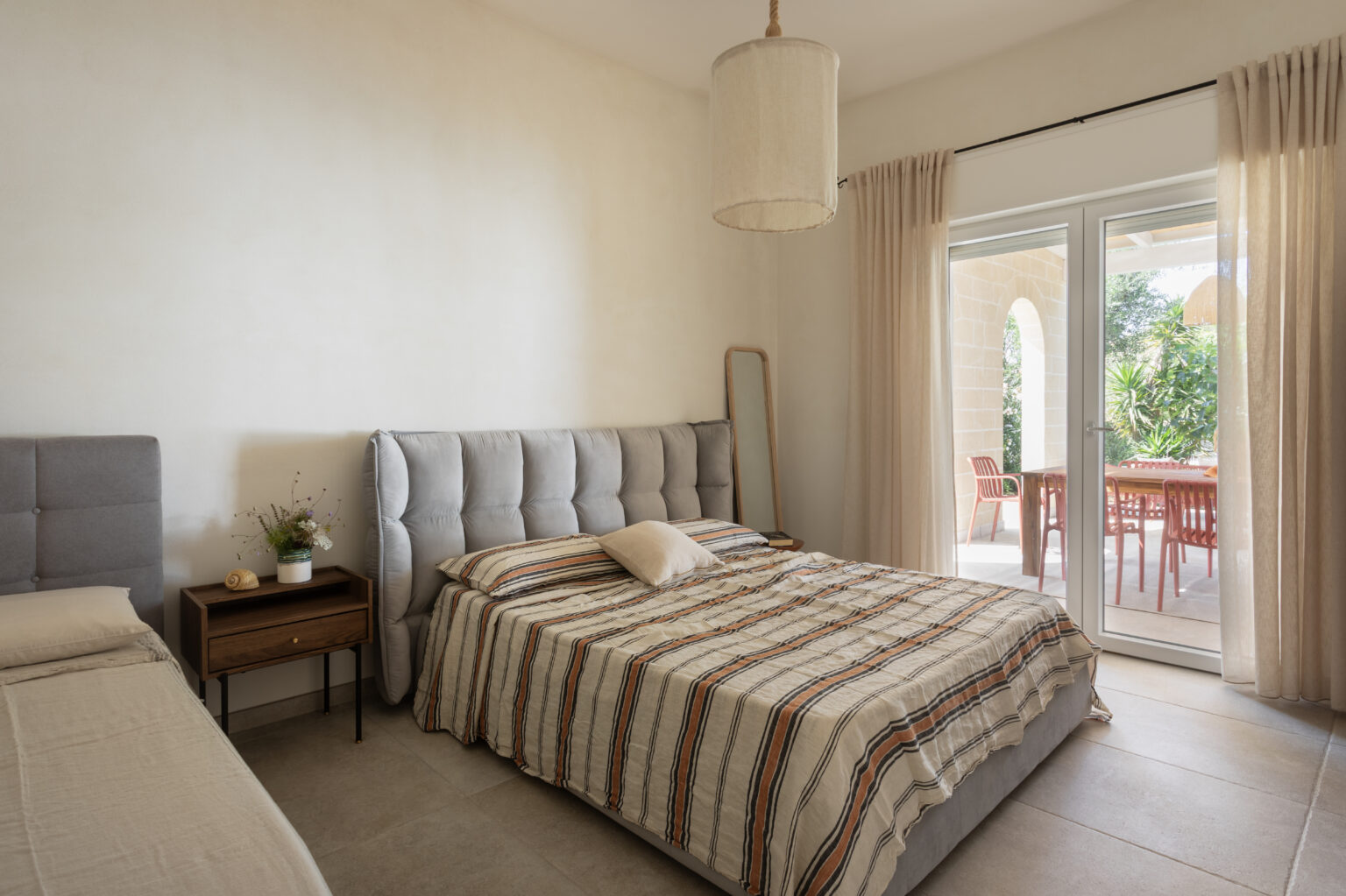 Modern bedroom featuring two stylish beds with tufted headboards, a bedside table with a decorative plant, and large glass doors leading to an outdoor patio area. The room is illuminated by natural light and has neutral-toned walls and soft curtains, creating a serene and inviting atmosphere. | Only on SopranoVillas