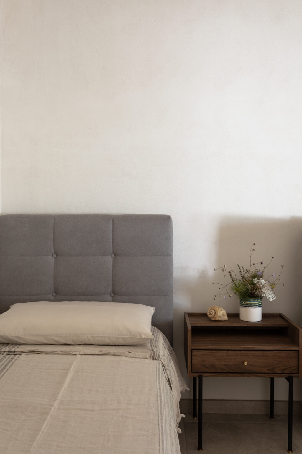 Modern bedroom interior featuring a gray upholstered headboard, a neutral-toned bedspread, and a minimalist nightstand with a small vase of flowers and a decorative shell. The soft color palette and clean lines create a serene atmosphere. | Only on SopranoVillas