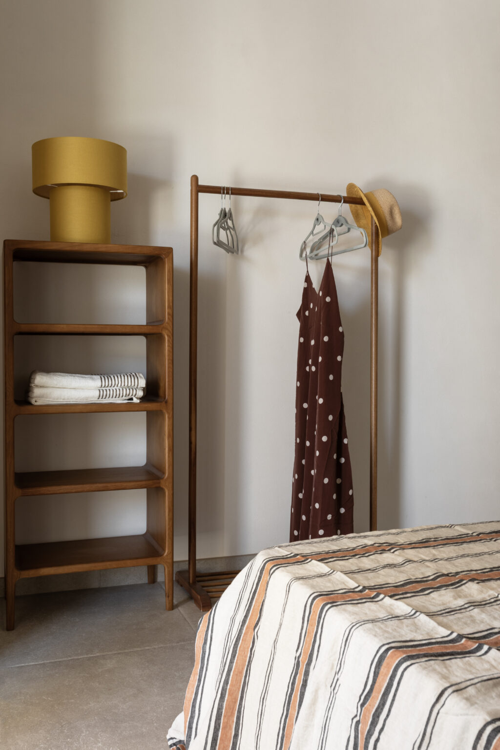 Cozy bedroom featuring a wooden clothing rack with a brown polka dot dress and a straw hat, complemented by a wooden shelf displaying neatly folded towels. A stylish yellow lamp adds a pop of color to the serene decor, enhancing the inviting atmosphere. | Only on SopranoVillas