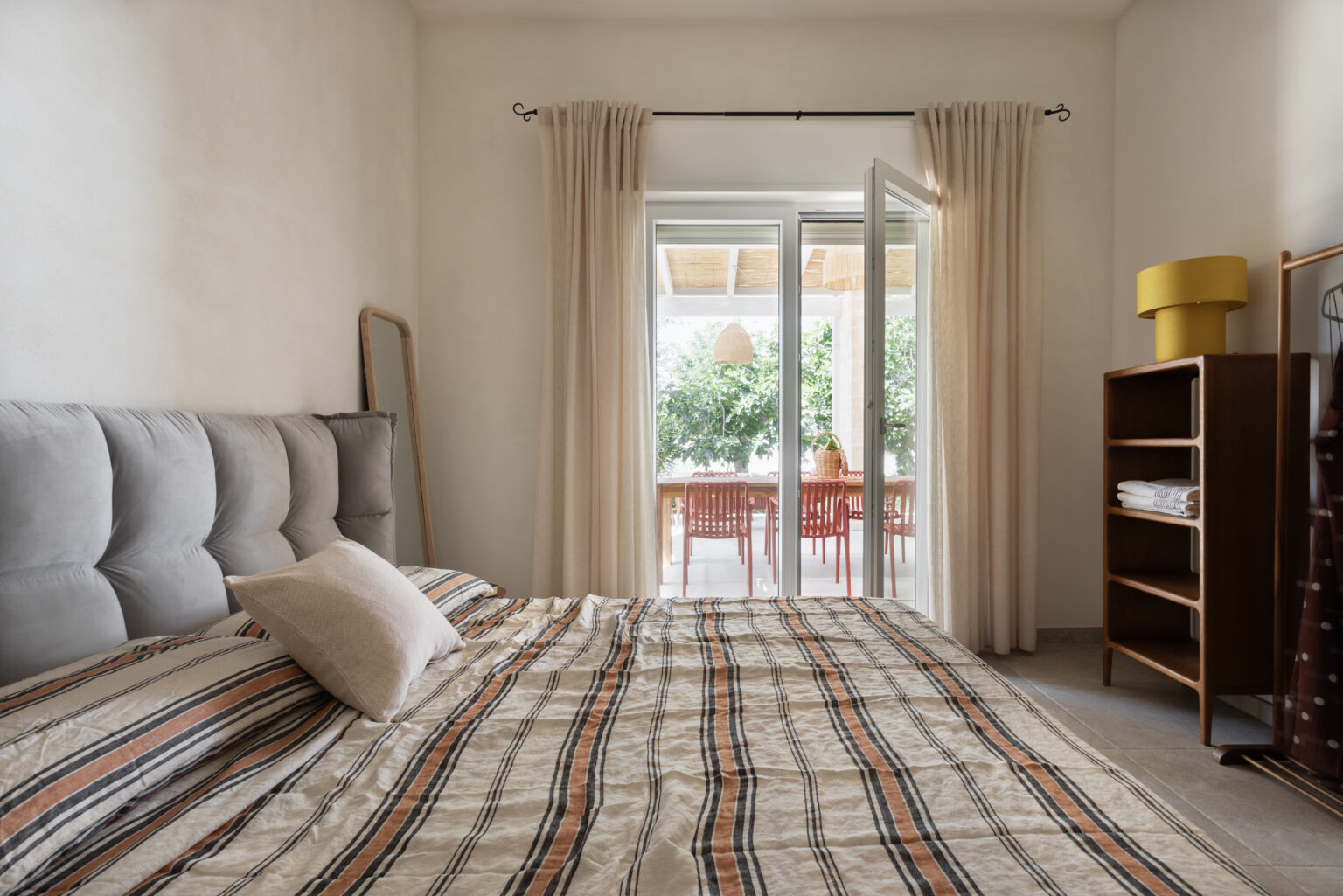 Modern bedroom interior featuring a comfortable gray upholstered bed with striped bedding, a decorative pillow, and a large window with sheer curtains leading to an outdoor dining area. A wooden shelving unit displays neatly folded towels, and a full-length mirror is positioned beside the bed, enhancing the spacious feel of the room. | Only on SopranoVillas