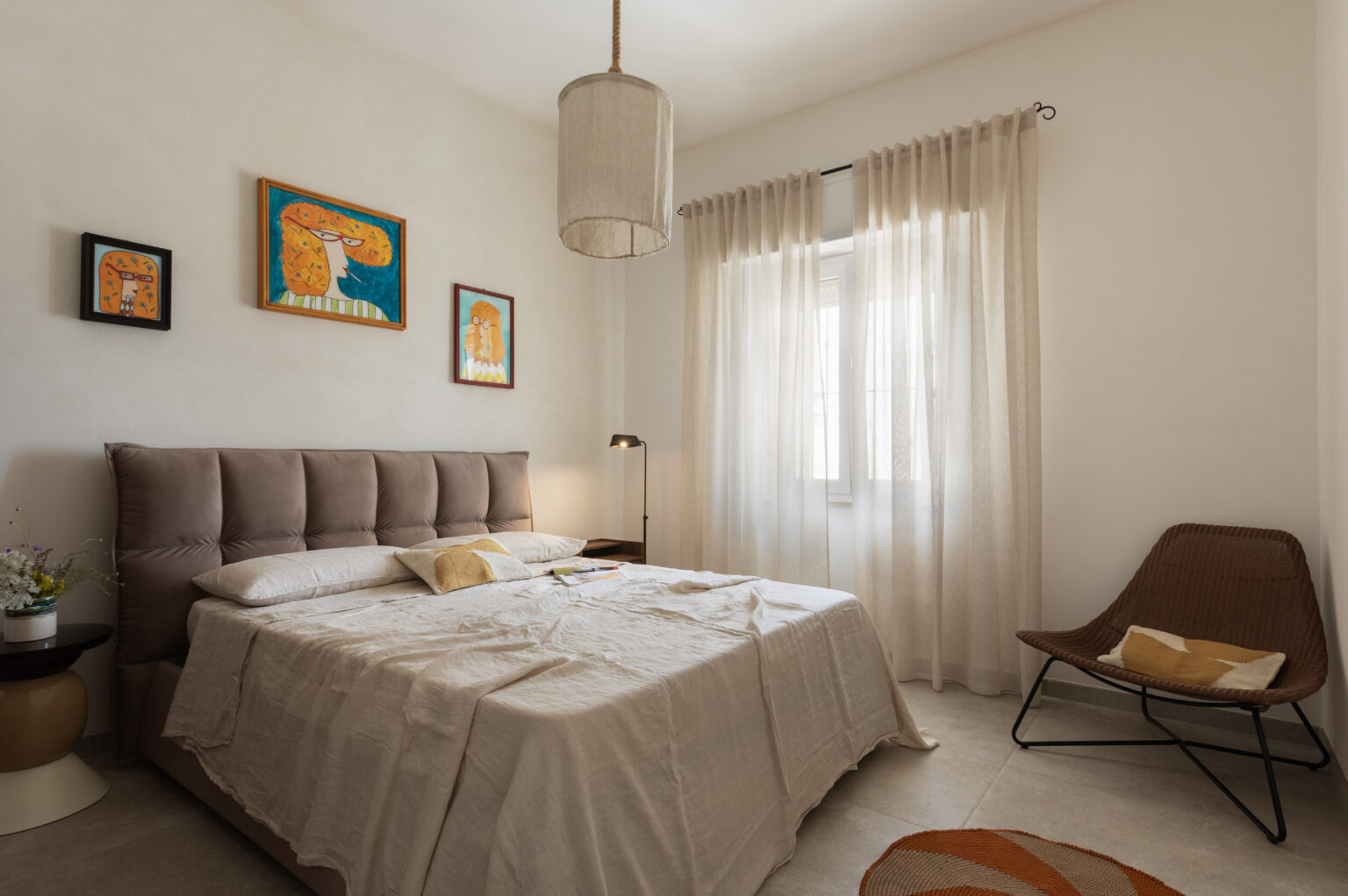 Cozy bedroom featuring a plush beige bed with a textured headboard, complemented by light-colored bedding and decorative cushions. The room is adorned with colorful artwork on the walls, a modern floor lamp beside the bed, and sheer curtains allowing natural light to filter in. A stylish woven chair and a round area rug add warmth and character to the space. | Only on SopranoVillas