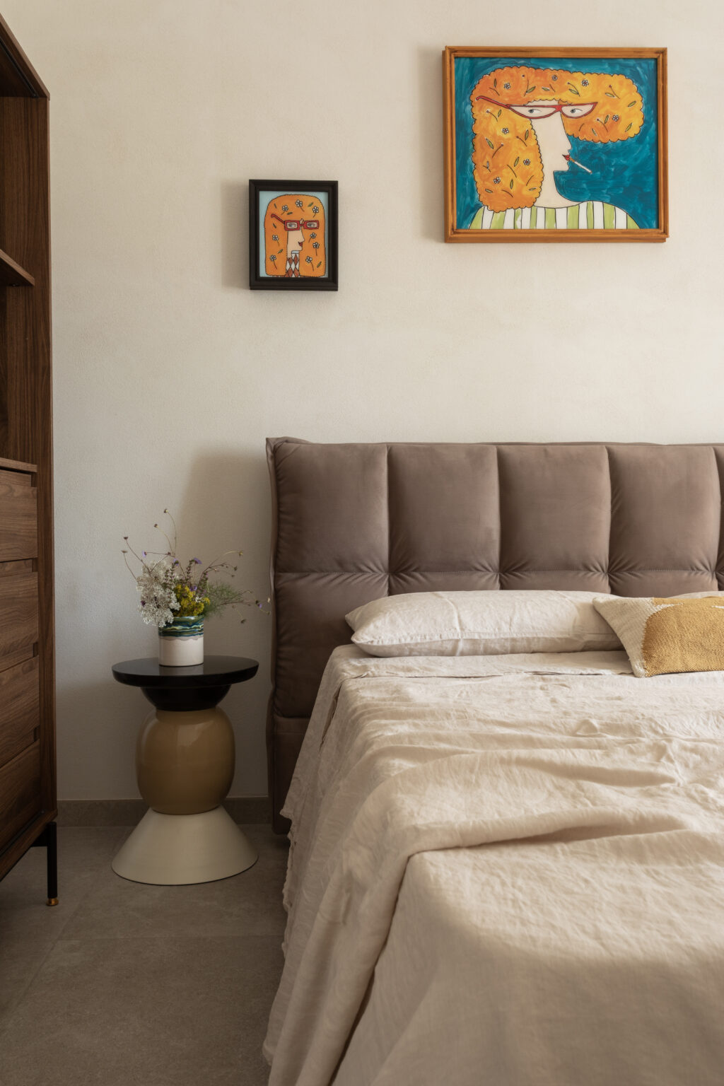 Cozy bedroom featuring a plush headboard, a neatly made bed with soft linens, and decorative pillows. Artwork adorns the walls, including two colorful paintings. A stylish side table holds a small vase with fresh flowers, enhancing the room's inviting atmosphere. | Only on SopranoVillas
