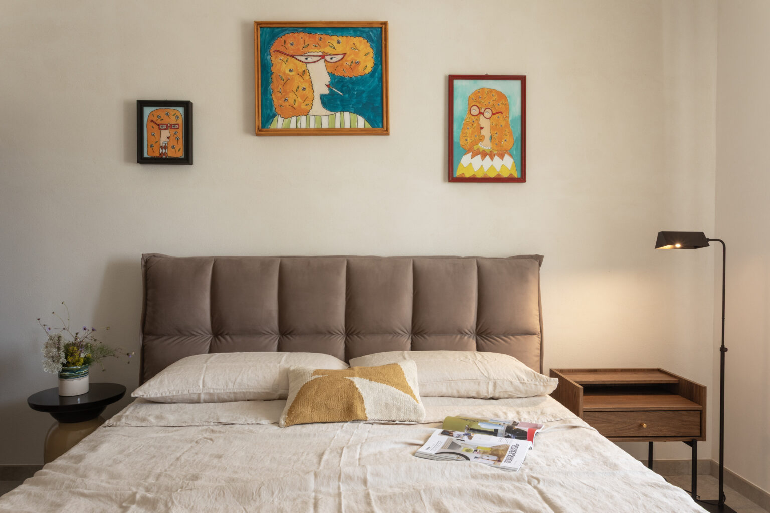 Cozy bedroom featuring a plush taupe headboard, decorative pillows, and an elegant bedside table with a lamp. The walls are adorned with colorful abstract paintings, and a small vase of wildflowers adds a touch of nature to the space. | Only on SopranoVillas