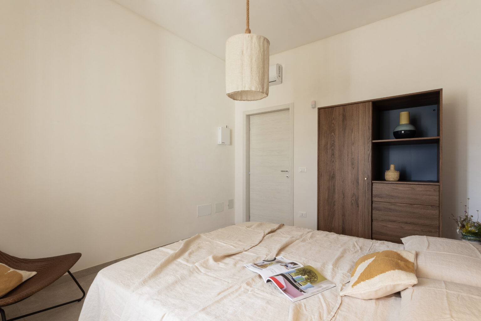 Cozy bedroom interior featuring a neatly made bed with a beige blanket, decorative pillow, and a magazine on top. A stylish wooden cabinet and a contemporary hanging light fixture add to the modern decor, complemented by soft neutral wall colors. | Only on SopranoVillas