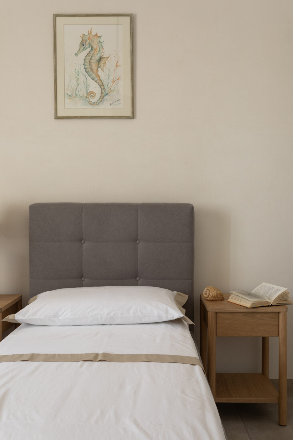 Cozy bedroom setup featuring a gray upholstered headboard, white bedding with a beige accent, and a wooden nightstand. The nightstand holds a seashell and an open book, while a framed watercolor seahorse hangs on the wall, adding a touch of nautical decor. | Only on SopranoVillas