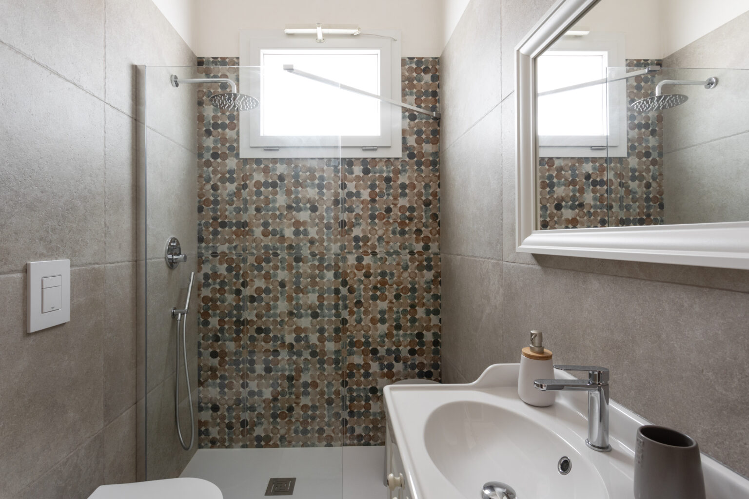 Modern bathroom featuring a glass shower with a rainfall showerhead, stylish mosaic tile wall, and a sleek sink with contemporary fixtures. Natural light streams in through a window, enhancing the clean and inviting atmosphere. | Only on SopranoVillas