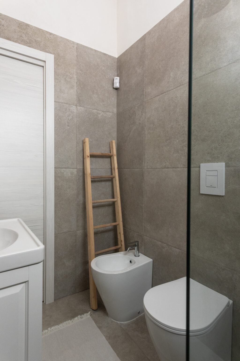 Modern bathroom interior featuring gray tiled walls, a white bidet, and a toilet, complemented by a wooden ladder and a glass shower enclosure. The design emphasizes a clean and minimalist aesthetic, ideal for contemporary home decor. | Only on SopranoVillas