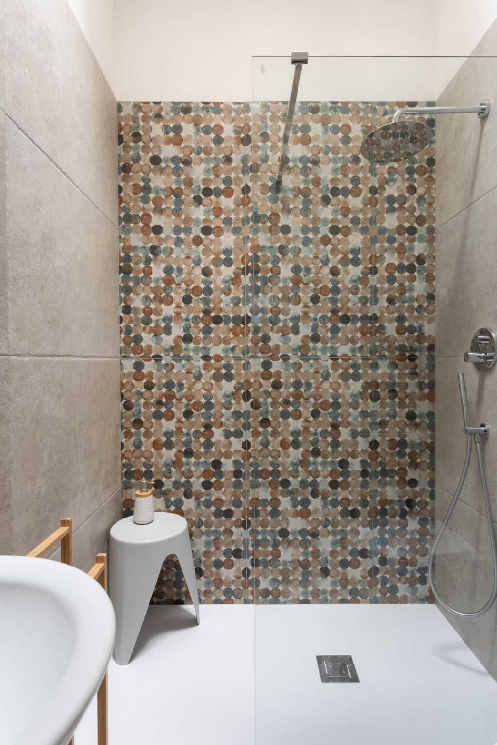 Modern shower design featuring a stylish glass enclosure, decorative circular tile pattern in earthy tones, and a sleek, minimalist stool. The space showcases contemporary bathroom aesthetics with neutral wall tiles and a functional layout. | Only on SopranoVillas
