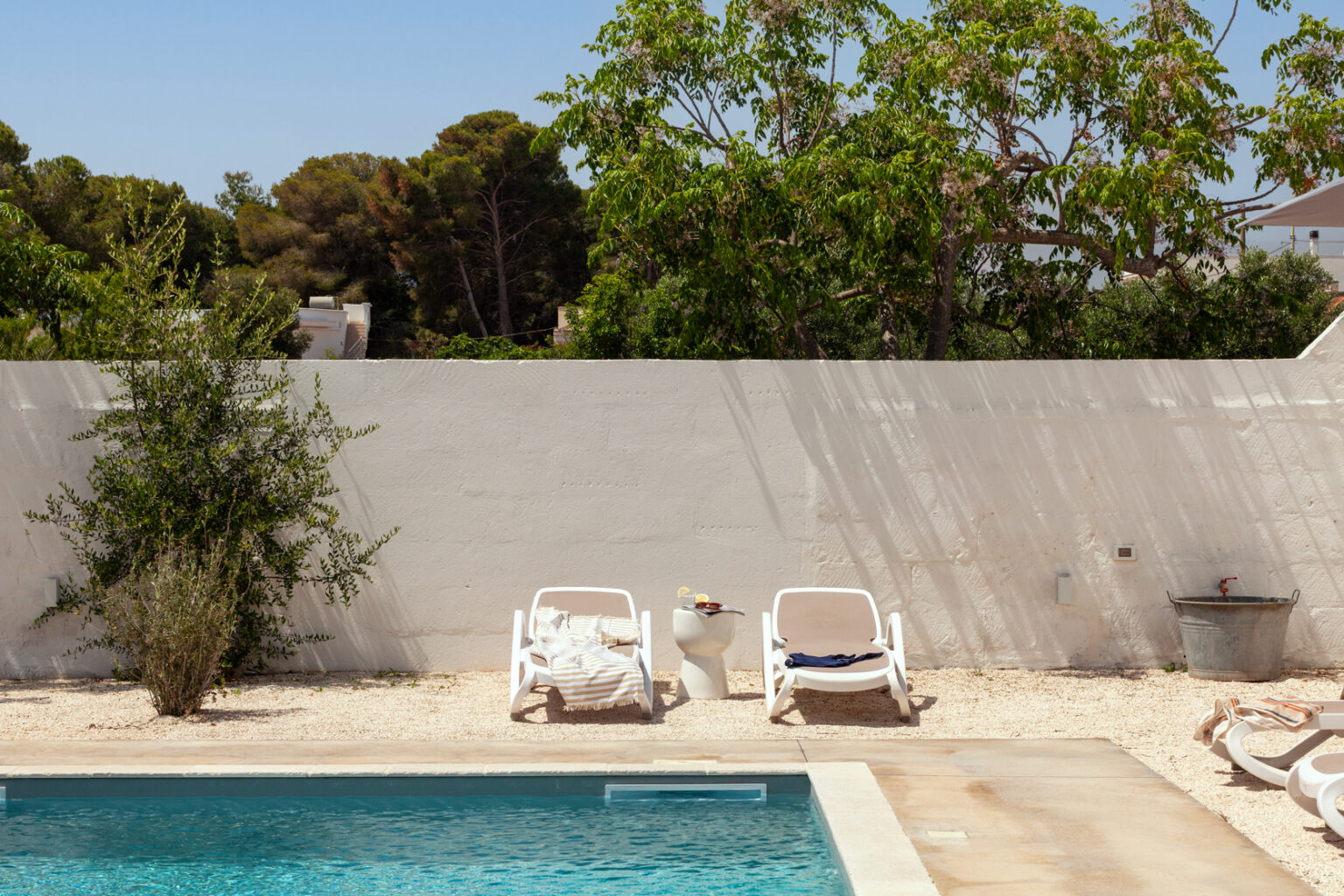 Relaxing outdoor pool area featuring two white lounge chairs, a small table with refreshments, and lush greenery in the background. The serene setting is perfect for sunbathing and enjoying a tranquil summer day. | Only on SopranoVillas