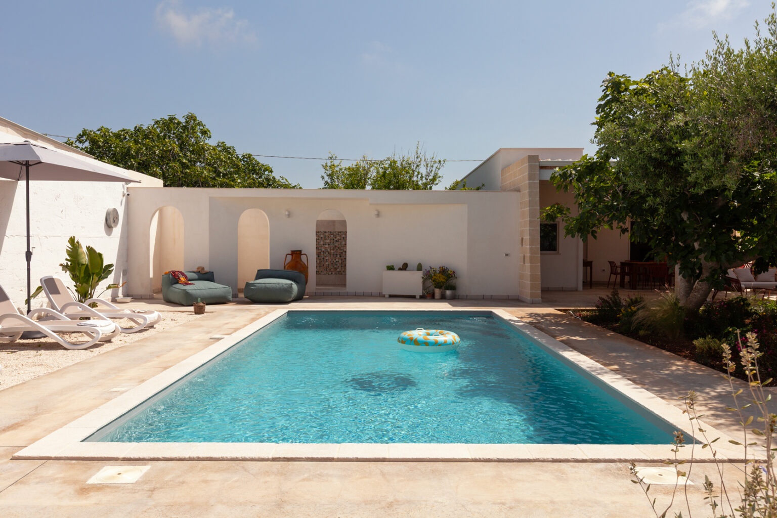 Modern outdoor pool area featuring a clear blue swimming pool, lounge chairs, and cozy seating. Surrounded by lush greenery and a stylish white wall, the space offers a relaxing atmosphere perfect for leisure and entertainment. | Only on SopranoVillas