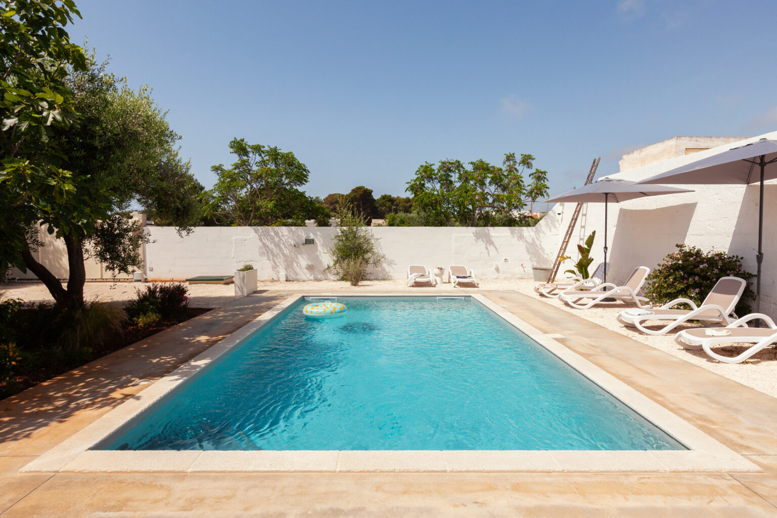 Swimming pool surrounded by lounge chairs and umbrellas in a serene outdoor setting, featuring lush greenery and a clear blue sky. Perfect for relaxation and leisure activities. | Only on SopranoVillas