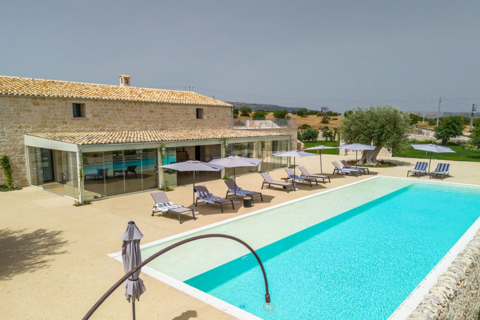 Contemporary villa with a spacious outdoor pool, lounge chairs, and umbrellas surrounded by lush greenery and a scenic landscape. Ideal for relaxation and vacation getaways. | Only on SopranoVillas
