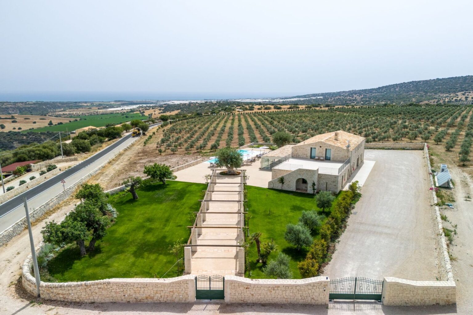 Aerial view of a spacious villa with a swimming pool, surrounded by lush green lawns and olive groves, overlooking a scenic landscape with distant hills and the sea. The property features a paved pathway leading up to the villa, bordered by landscaped greenery, providing a tranquil atmosphere in a rural setting. | Only on SopranoVillas