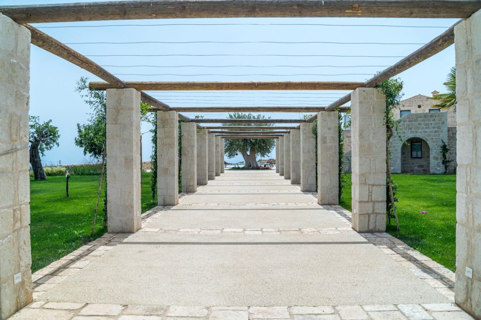 Pathway lined with stone columns and wooden beams, leading through a lush green garden with trees and seating areas, against a clear blue sky. Perfect for outdoor relaxation and scenic strolls. | Only on SopranoVillas