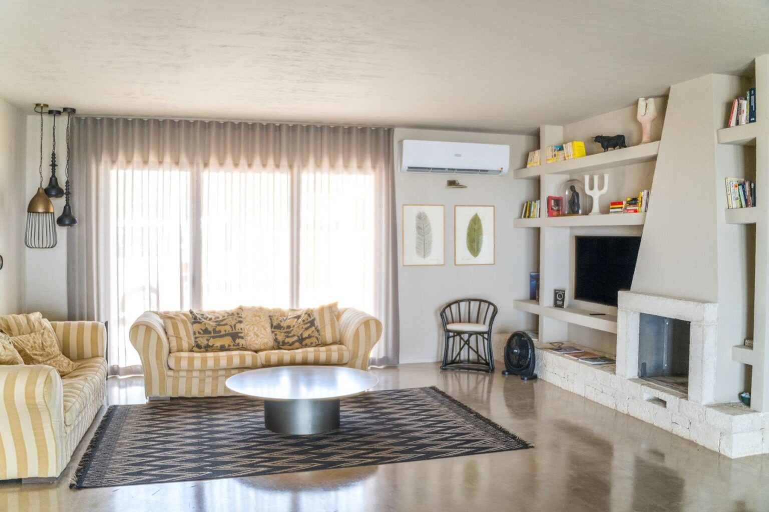 Modern living room featuring striped sofas, a round coffee table, and a decorative rug. Large windows with sheer curtains allow natural light. Shelves displaying books and decorative items line the wall, alongside a minimalist fireplace and air conditioning unit. | Only on SopranoVillas